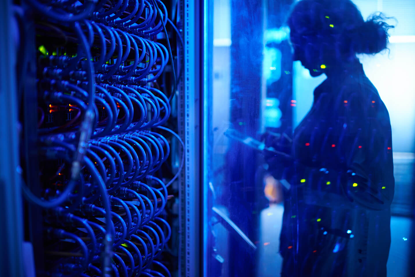 Technikerin mit Tablet in blau beleuchtetem Serverraum, Netzwerkverkabelung und Lichtern im Hintergrund sichtbar